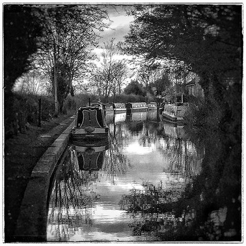 A romantic black and white photograph of narrowboats lining the canal with trees overhead